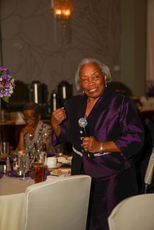 A speaker in a purple satin jacket addresses dinner guests at an elegant event with white tablecloths.
