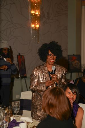 A speaker in a shimmering gold outfit addresses guests at a formal dinner event with elegant chandeliers overhead.