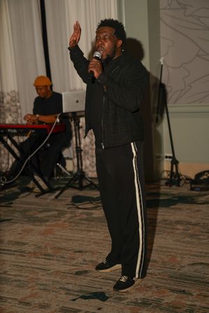 A performer in athletic wear sings on stage with a keyboard player visible in the background.
