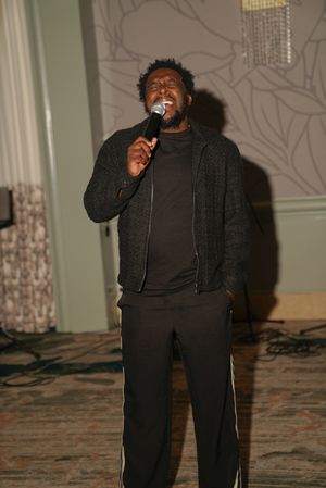 A performer in all black attire holds a microphone while singing on stage with decorative wall patterns behind them.