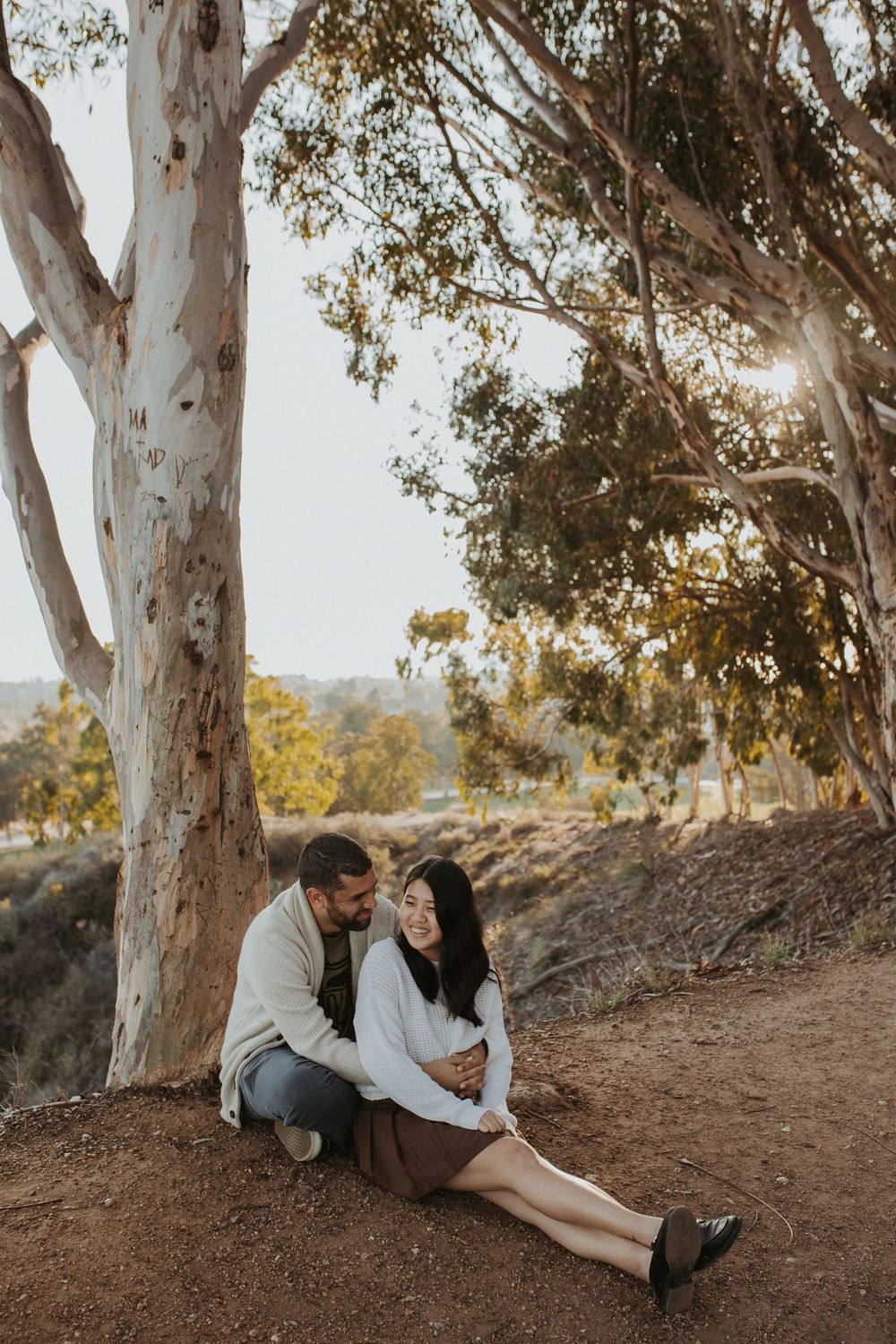 A man and woman sitting on the ground smiling at each other at sunset.