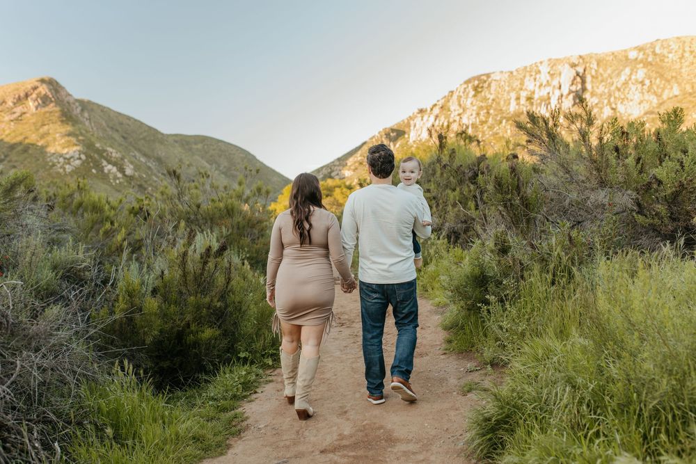 A man, woman, and baby walking away with mountains and grass in the background.