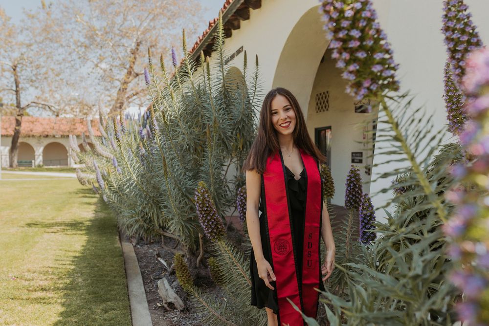 A woman wearing a red graduation sash standing between archways and tall purple flowers.
