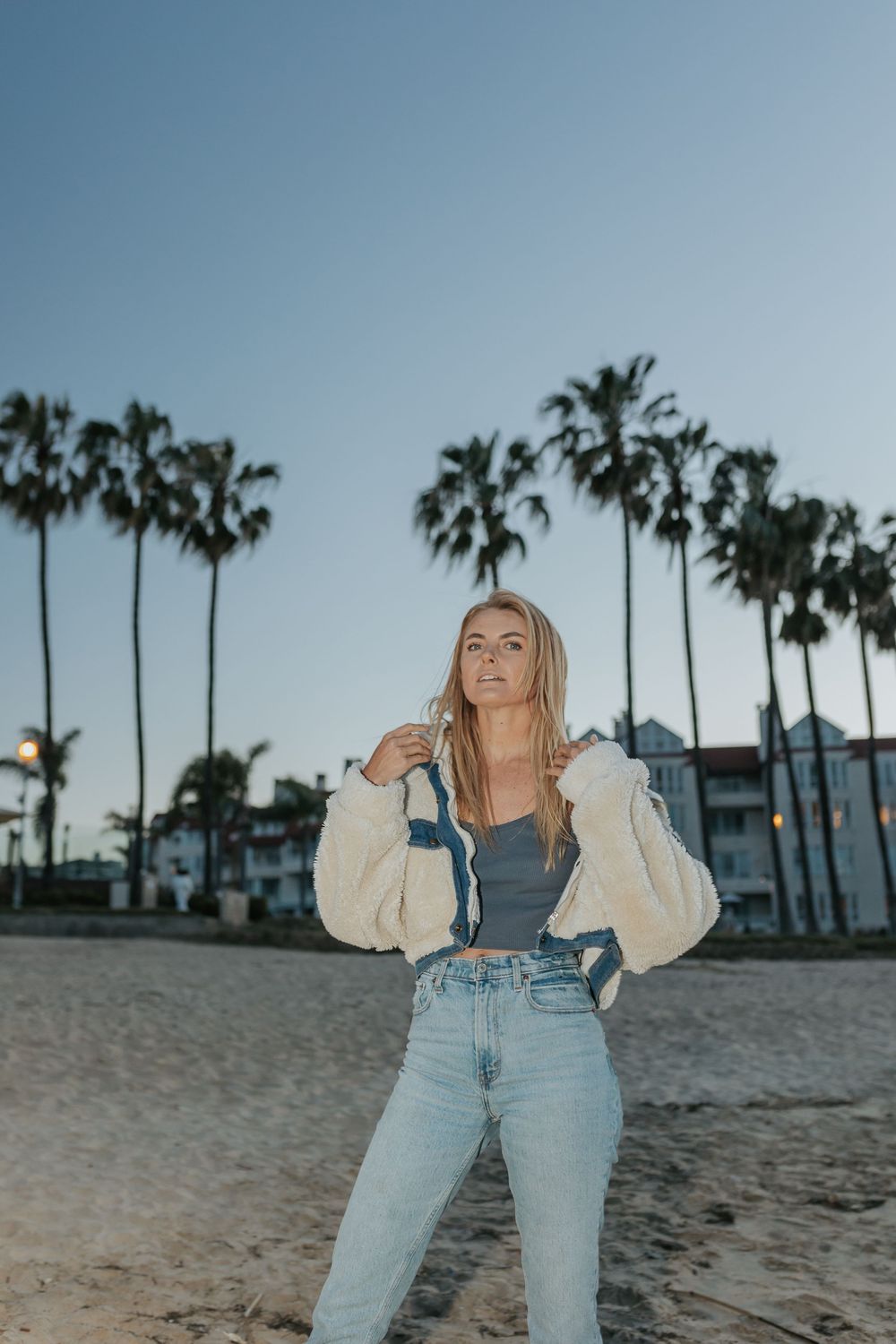A blonde woman stands on a beach with palm trees behind her at night.