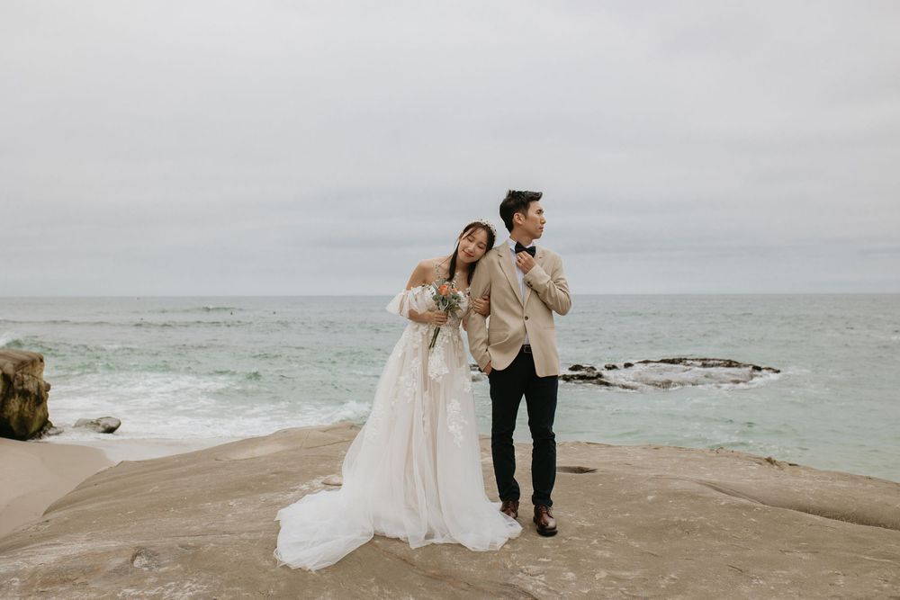 A man and woman in wedding attire on a cloudy day with the ocean behind them.