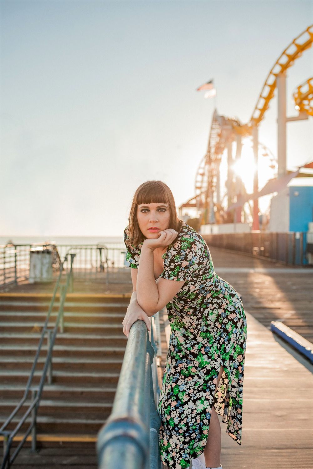 A woman wearing a green floral dress leans on a railing at the Santa Monica Pier.