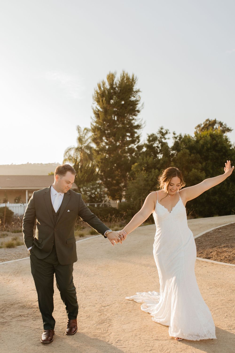 A man and woman in wedding attire holding hands, walking, and smiling during a sunset.