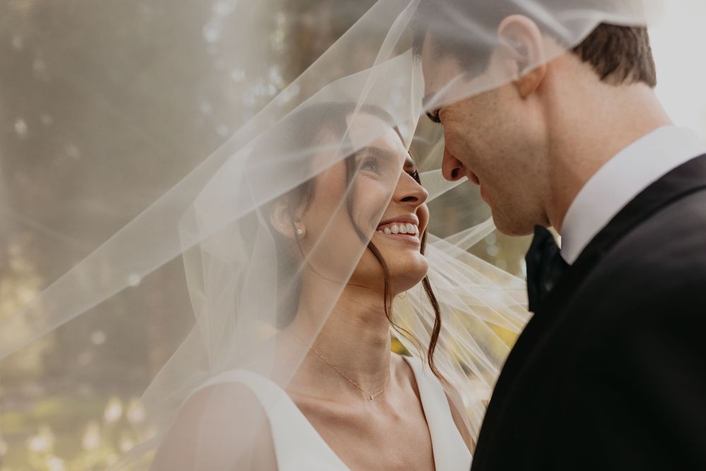 A man and woman in wedding attire face each other while under the bride's veil.