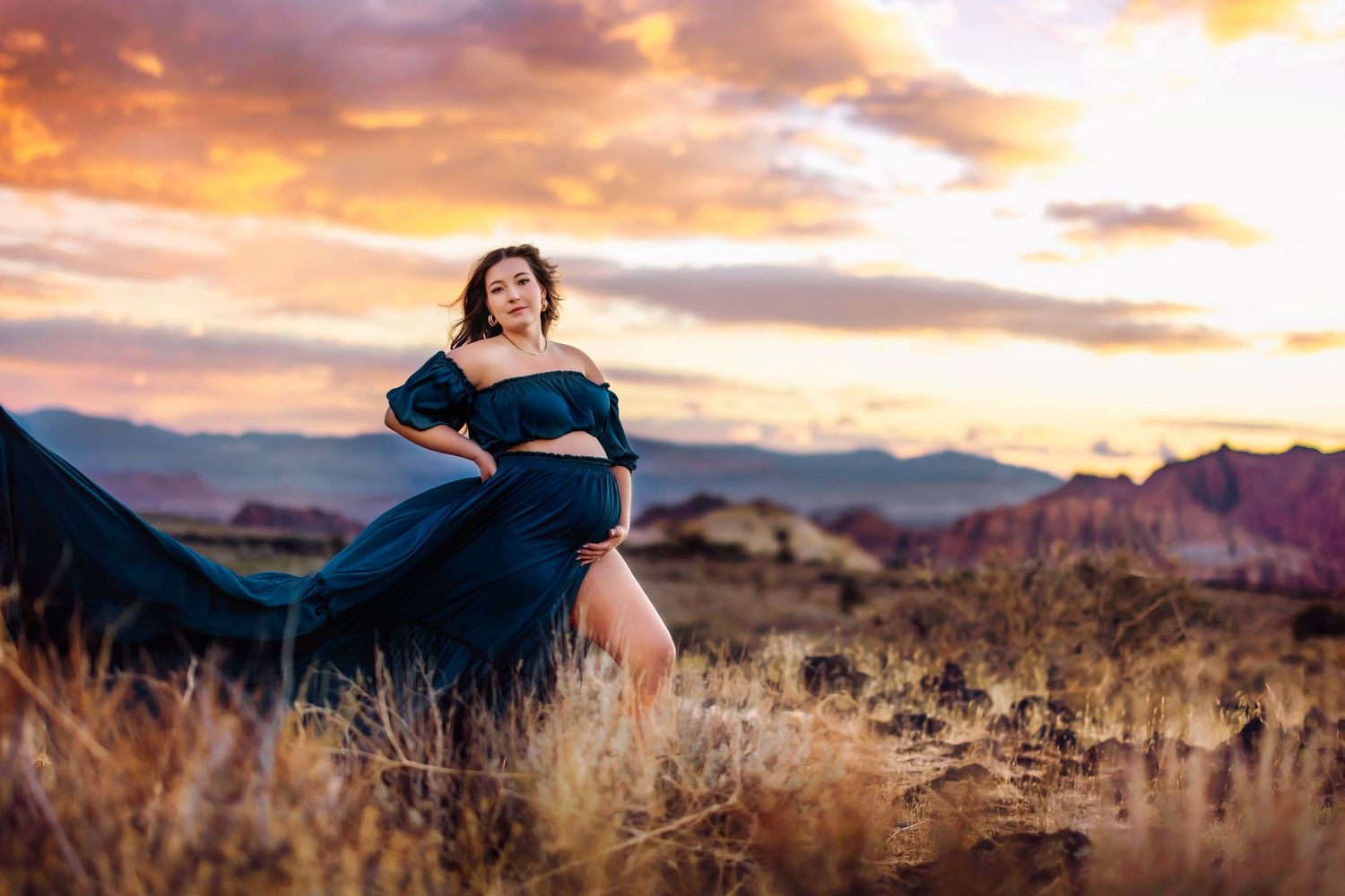 A maternity photo session in a flowing blue dress against a dramatic desert sunset.