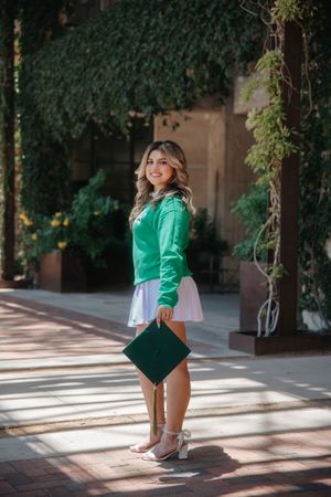 Graduate in green and white dress sits holding graduation cap in outdoor corridor with dappled sunlight.