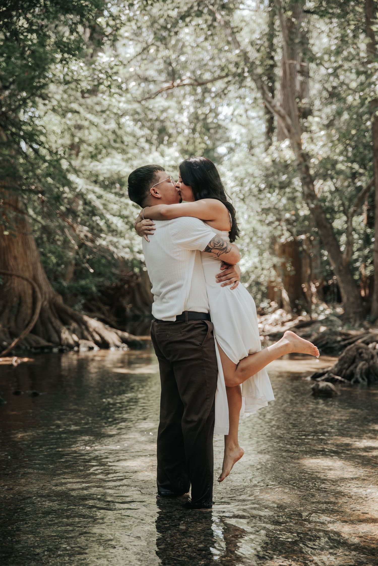A romantic couple embraces while standing in shallow creek waters surrounded by lush forest trees.