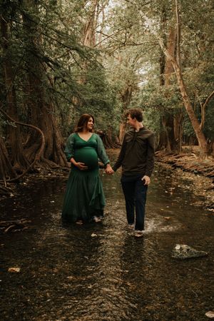 Couple holding hands during maternity shoot in lush forest creek setting.