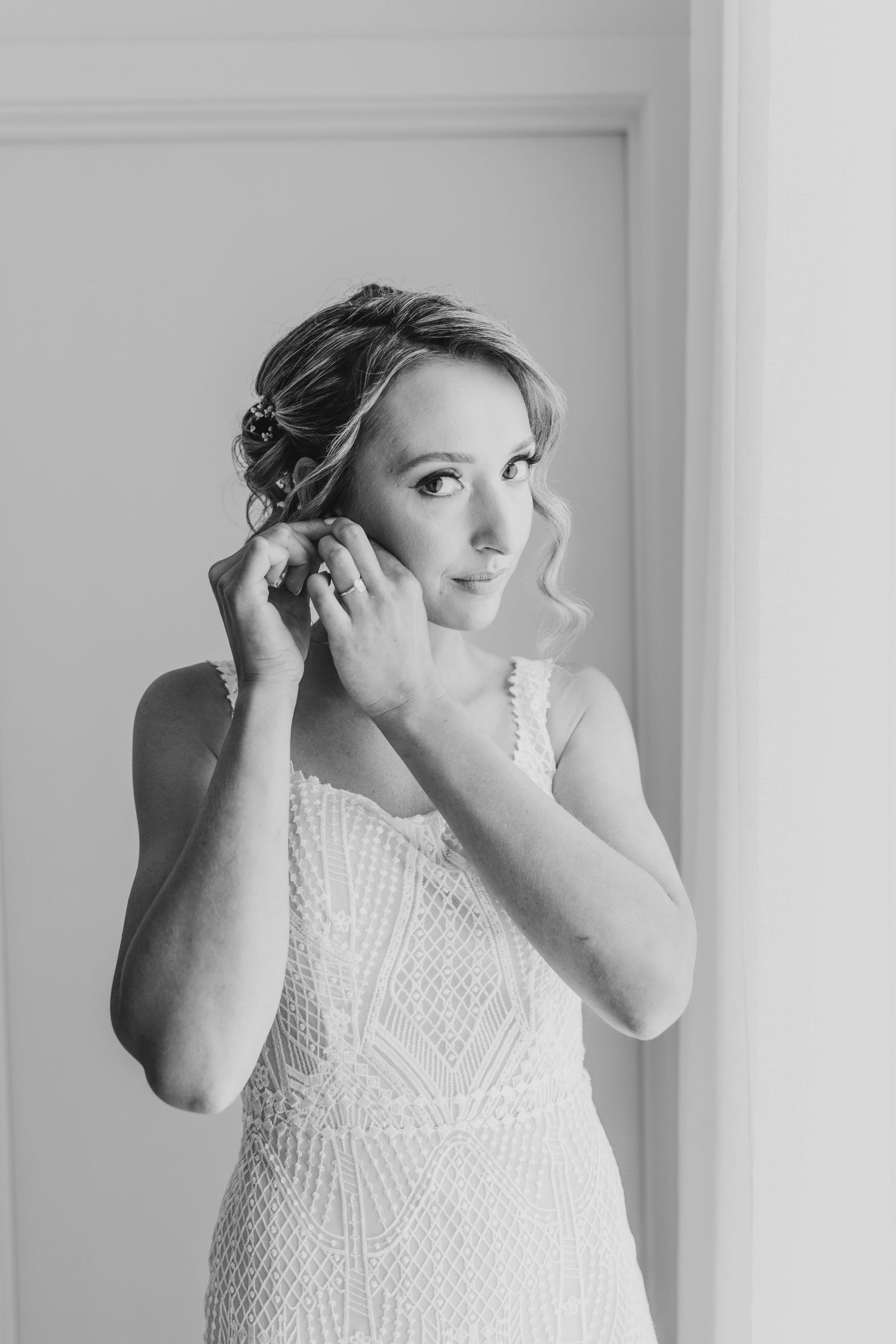 A bride in a detailed lace dress adjusts her earring in a black and white portrait.