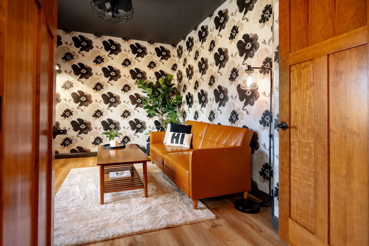 Cozy sitting room with bold floral wallpaper, tan leather sofa, wood coffee table, and plush white rug on hardwood floors.