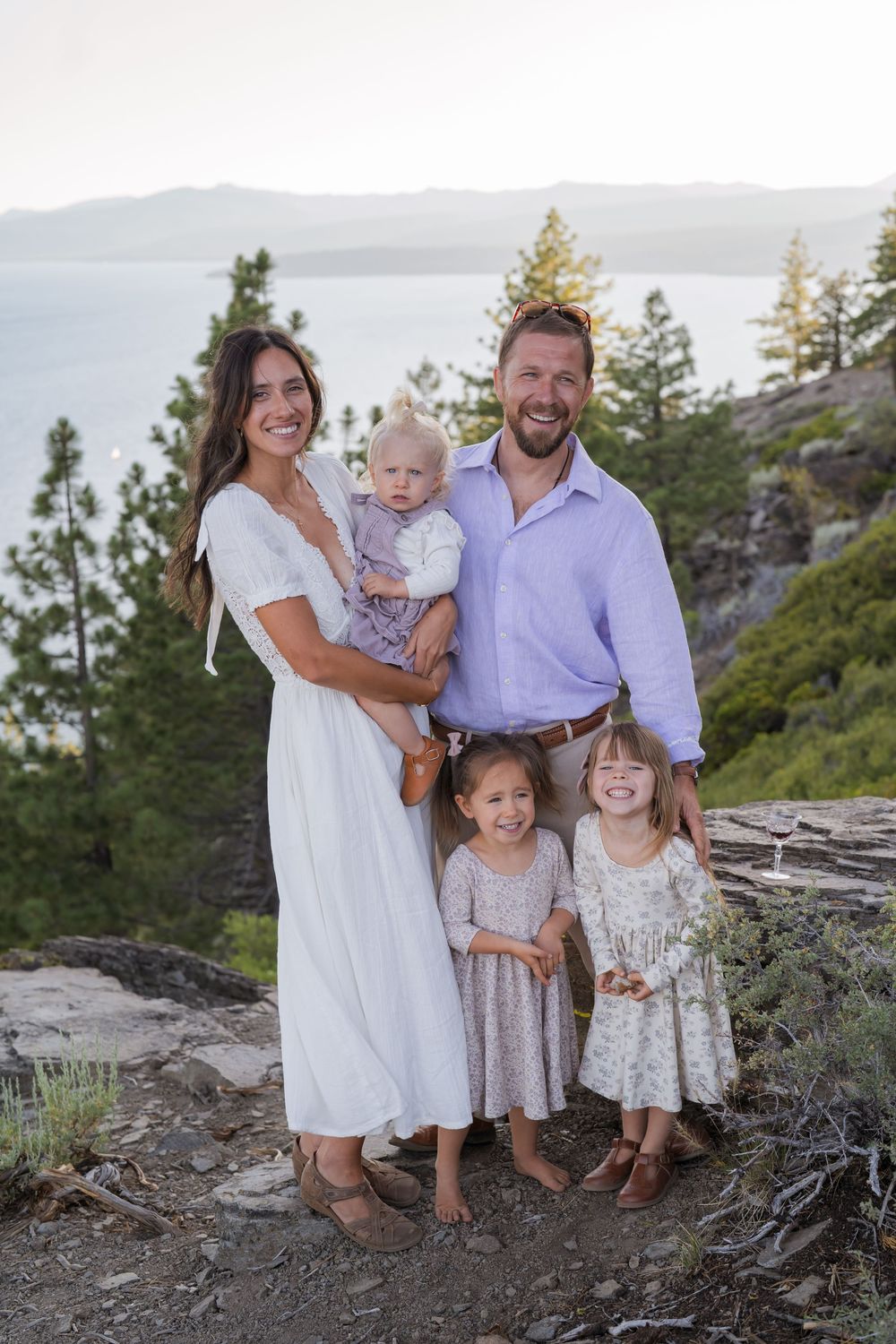 Family wearing white and floral dresses poses together on rocky mountain overlook during sunset photoshoot.