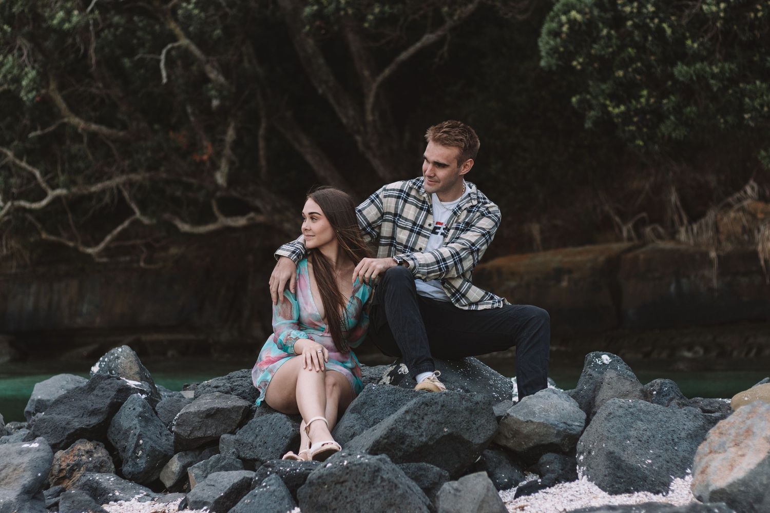 A couple sits together on rocky shoreline wearing casual clothes while enjoying a peaceful moment in nature.