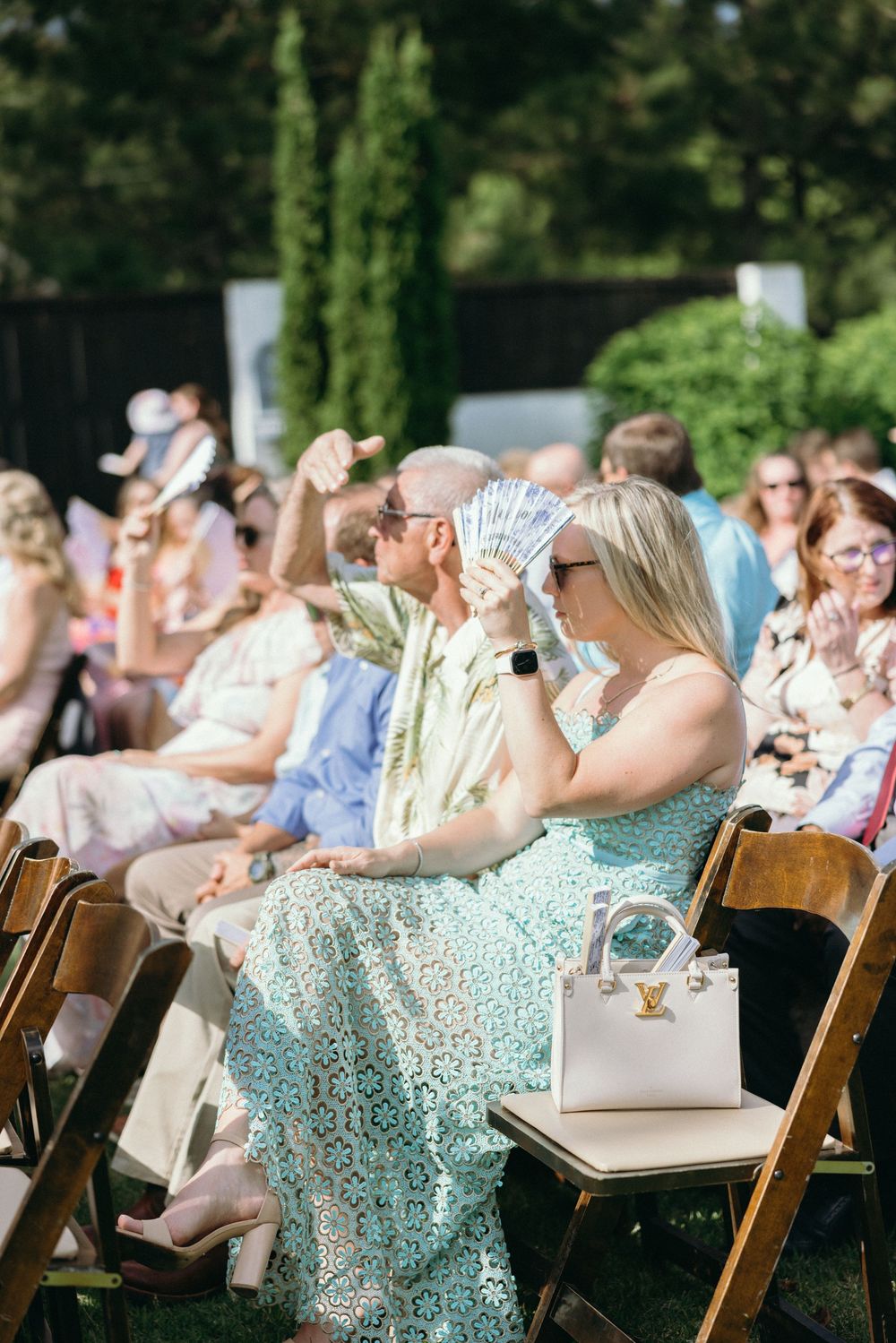 Wedding guests seated in wooden chairs fan themselves during an outdoor ceremony. The Bradford, Separk  Mansion, Graylyn Estate