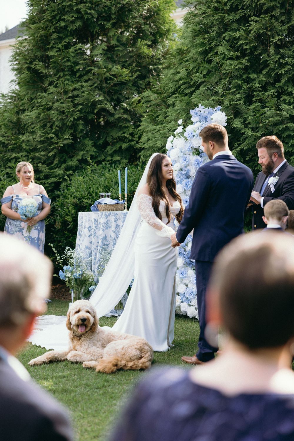 An outdoor wedding ceremony with blue floral decorations and guests seated on wooden chairs. The Bradford, Separk  Mansion, Graylyn Estate