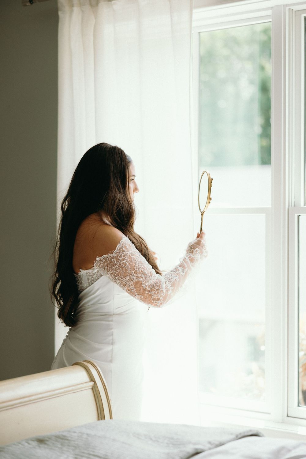 A bride in a white lace gown gazes out a bright window while holding a vintage hand mirror. The Bradford, Separk  Mansion, Graylyn Estate