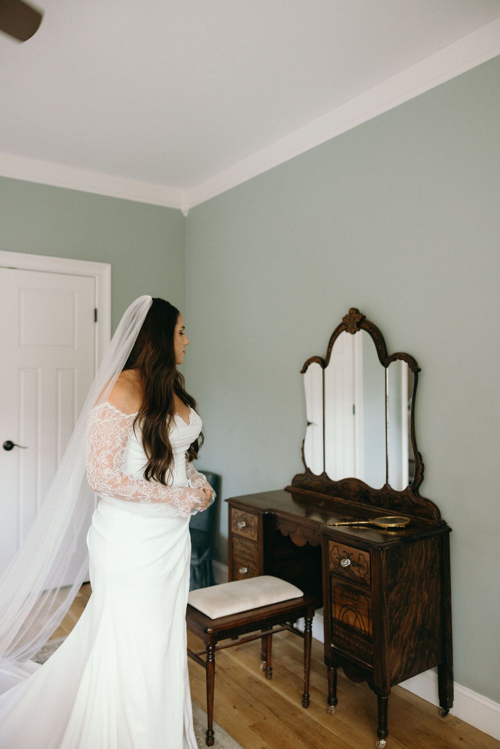 A bride in an elegant white lace dress stands before an antique vanity mirror in a sage green room. The Bradford, Separk  Mansion, Graylyn