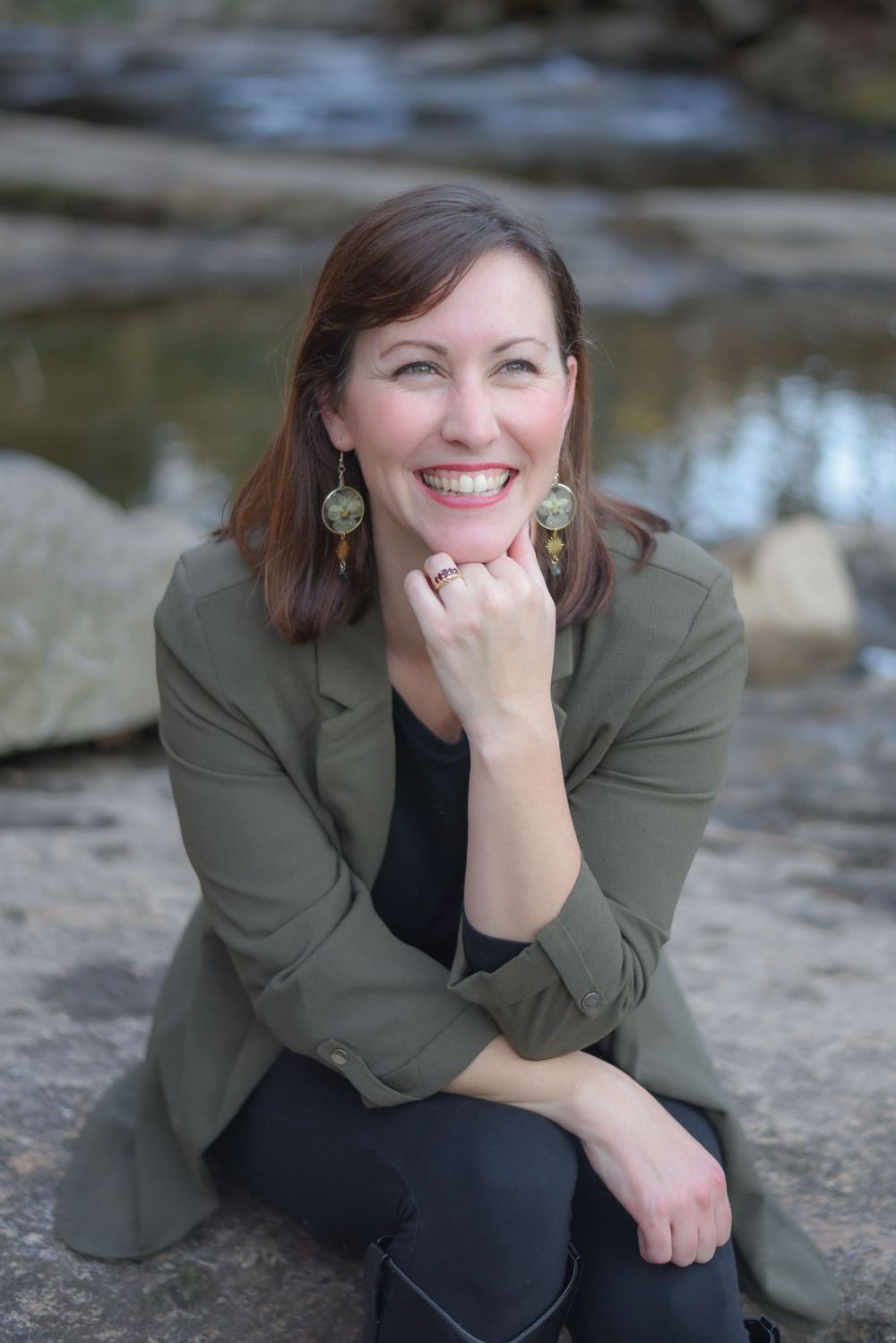 Professional headshots of person in olive blazer sitting on rocks near water.