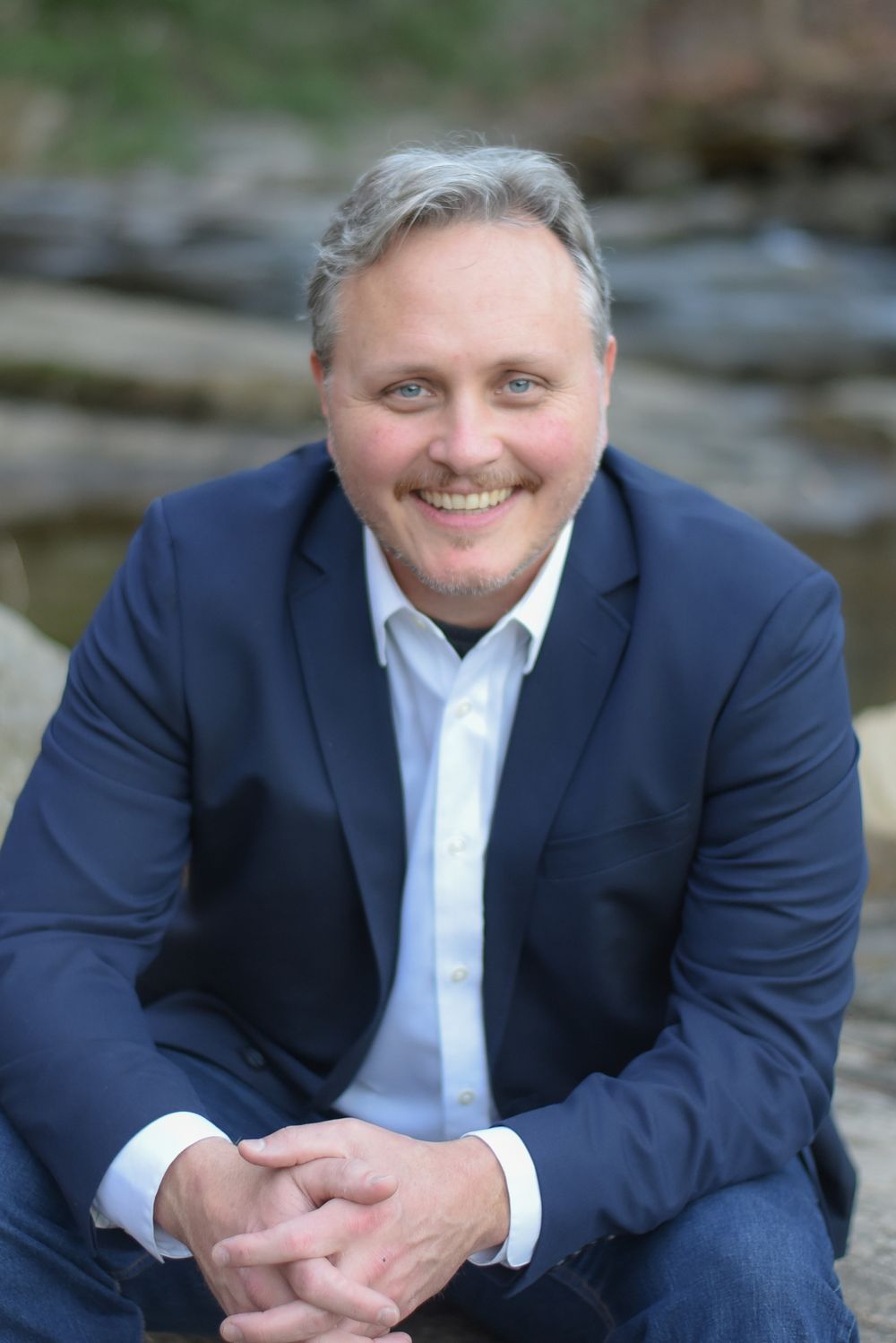 Professional headshot portrait of person in navy suit and white shirt against stone background.