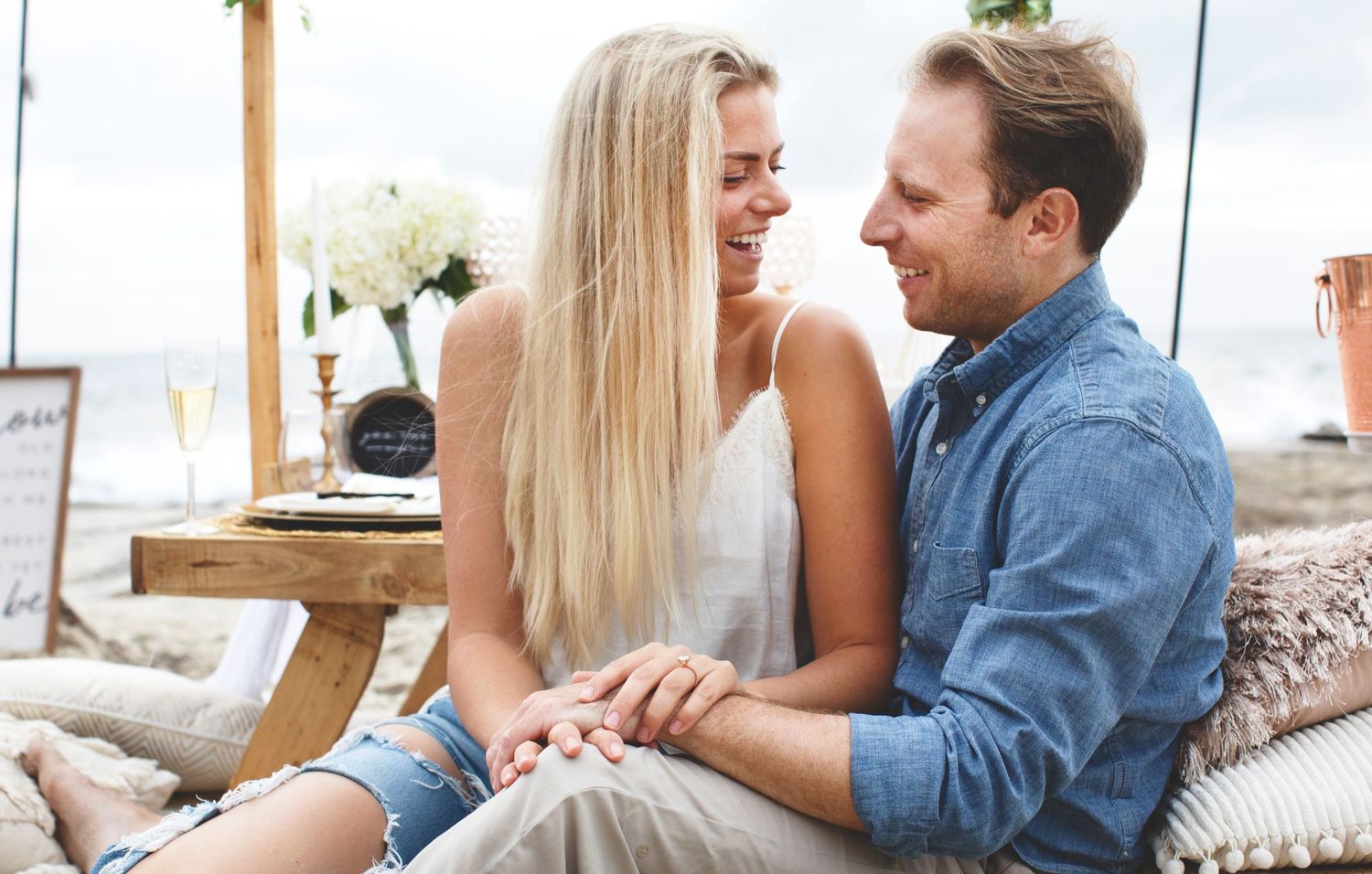 Couple sharing intimate moment at beach picnic with decorative setup and pillows.
