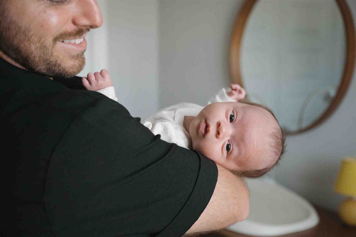 A smiling adult holds a wide-eyed baby wearing white clothing while standing in a bedroom interior.