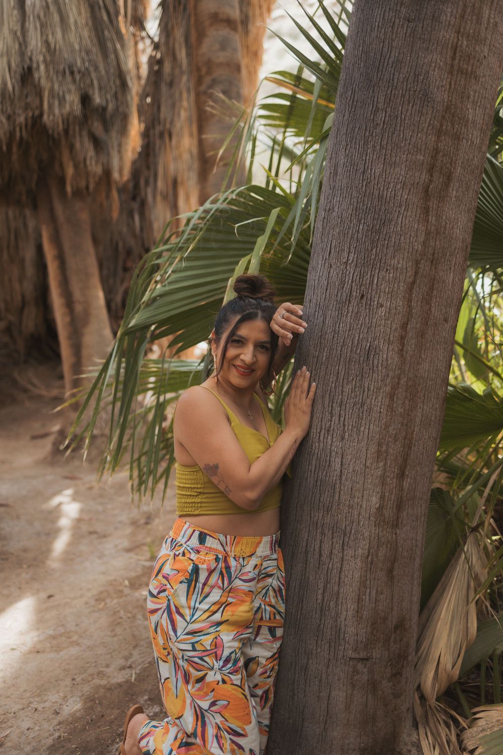 Someone in a yellow tank top and patterned pants poses against a palm tree trunk.