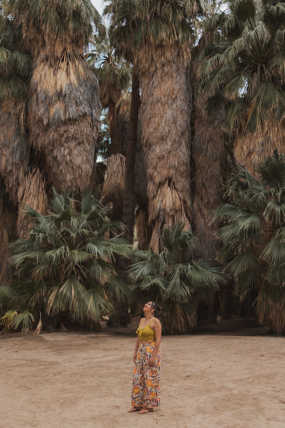 A figure in denim and yellow trousers poses against a weathered concrete structure in a desert setting.