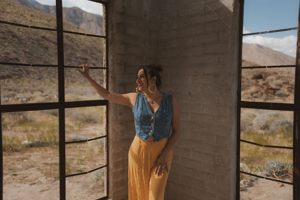 Someone stands in an abandoned desert building with large windows and mountain views.