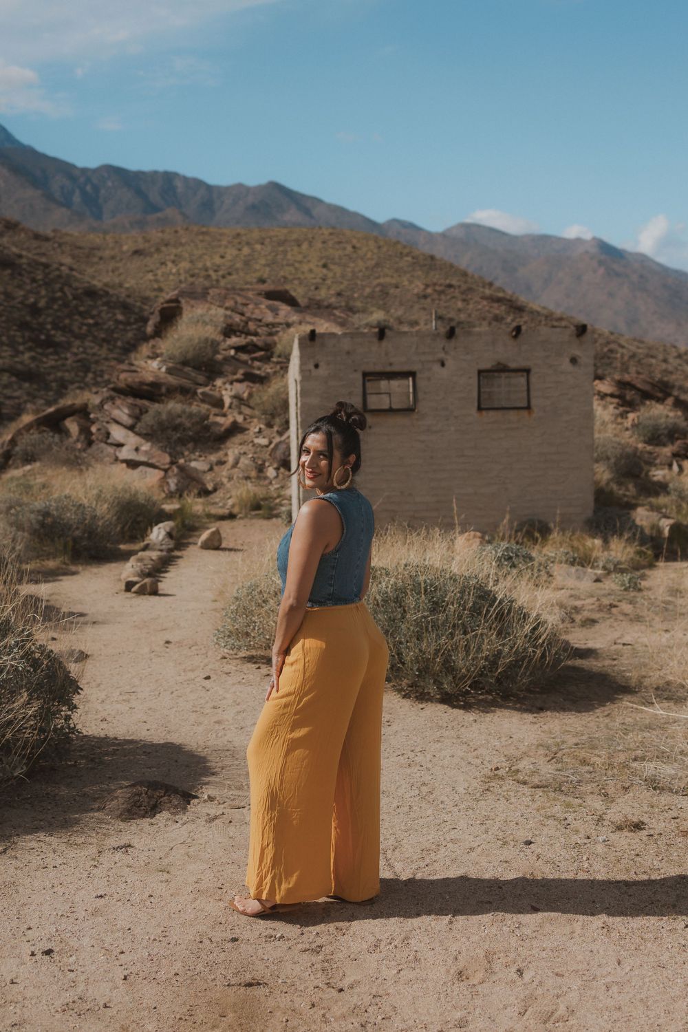 Person in yellow wide-leg pants and blue top poses in front of abandoned desert building with mountains in background.