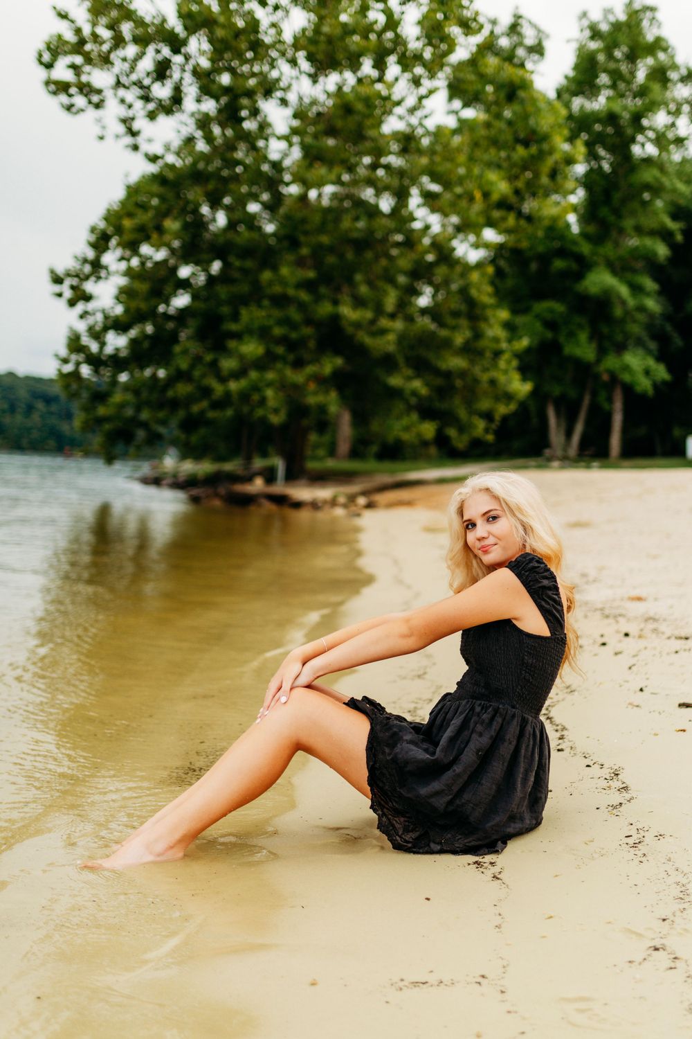 Blonde woman in black dress sits on sandy lakeside beach with green trees and calm water nearby.