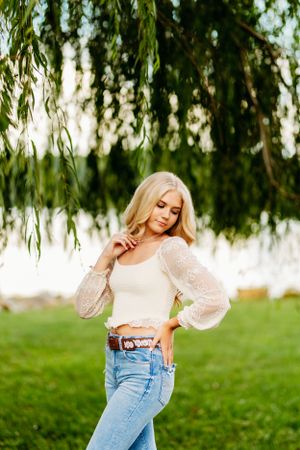 Blonde girl in white lace top and ripped jeans posing gracefully under a weeping willow tree.