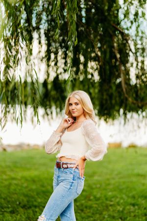 Blonde teenage girl in white crop top and jeans posing under a weeping willow tree by a lake.