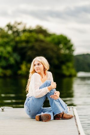 Blonde teen girl sitting on a wooden dock by a lake in jeans and white top for senior photos.
