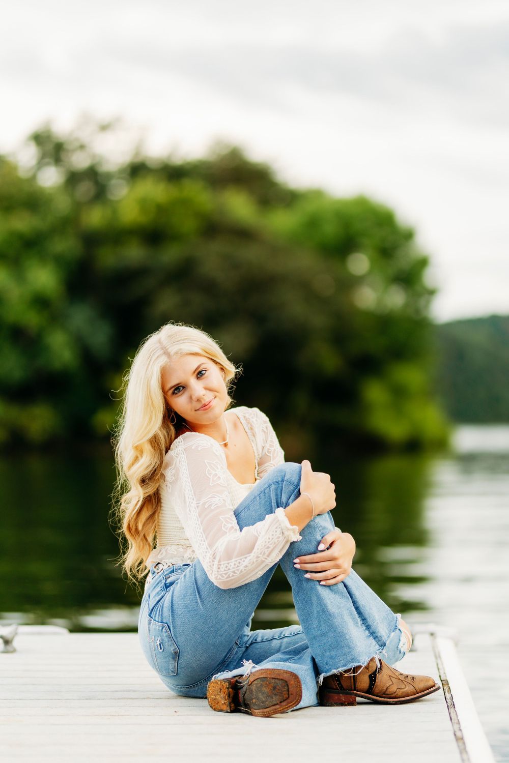 Smiling blonde girl with long wavy hair sitting on a lakeside dock in jeans and white lace top.