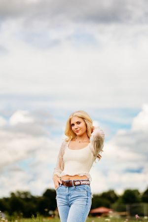 Blonde woman in white crop top and jeans posing in a flower field against a dramatic cloudy sky.
