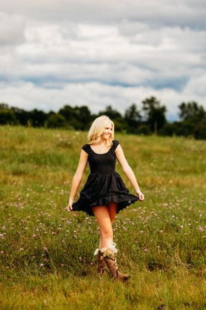 Blonde woman in black dress and cowboy boots twirling in a wildflower field under a dramatic sky.
