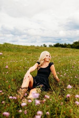 Blonde woman in black dress sitting in a wildflower meadow with purple flowers under a cloudy sky.
