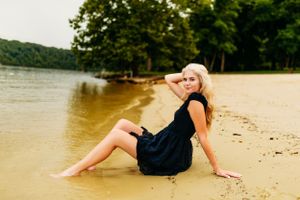 Blonde woman in black dress sits on sandy beach shore with calm lake and green trees behind her.