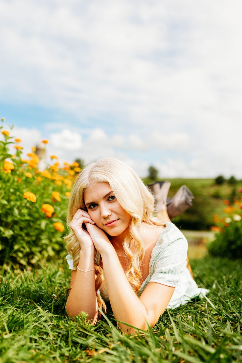 Blonde woman lying in green grass beside orange flowers, resting her face in her hands outdoors.