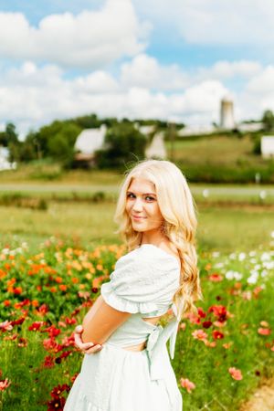 Smiling blonde woman in light blue off-shoulder dress posing in a flower field with a farm backdrop.