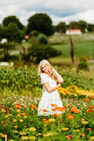 Young blonde woman in white dress standing in a vibrant flower field with orange and yellow blooms.