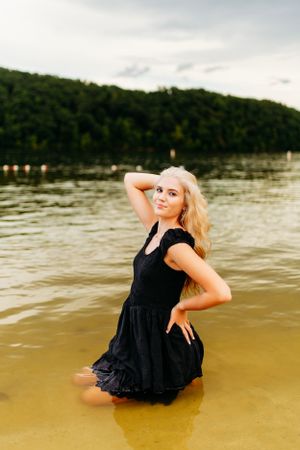 Blonde woman in black dress poses in lake water with lush green hillside and cloudy sky behind her.