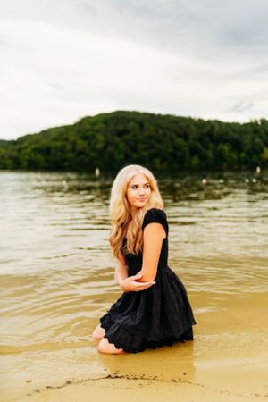 Blonde woman in black dress kneels in shallow lake water with green forested hills in background.