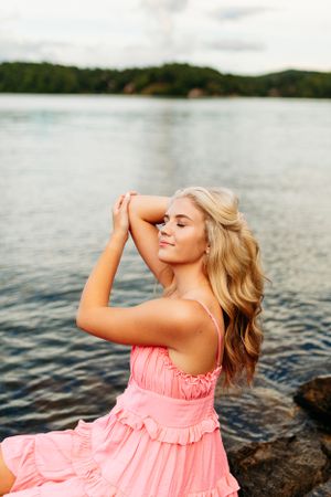Blonde girl in pink ruffled dress sitting on rocks by a lake with eyes closed for senior photos.
