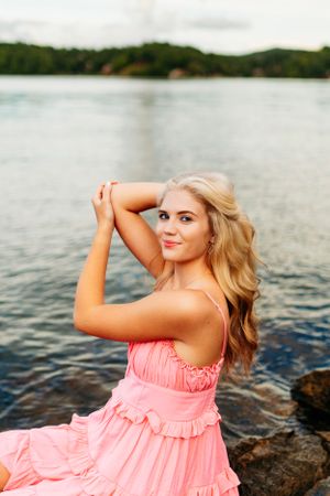 Smiling blonde girl in pink sundress posing on lakeside rocks with arms raised for senior photos.
