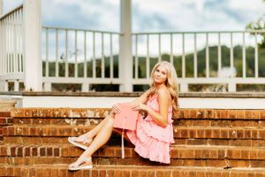 Blonde girl in pink sundress sitting on brick steps holding a pink clutch for senior portraits.