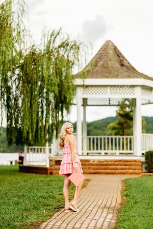 Blonde woman in pink dress poses on brick path near white gazebo by lake with weeping willow tree.